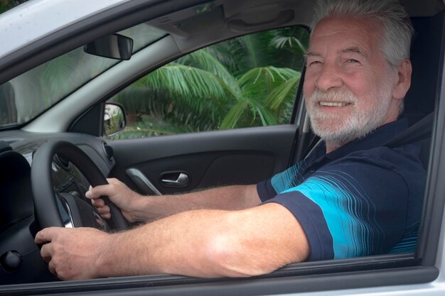 Un homme âgé attrayant sourit après avoir garé la voiture Personnes âgées caucasiennes aux cheveux gris et à la barbe Plantes vertes à l'extérieur de la fenêtre de la voiture