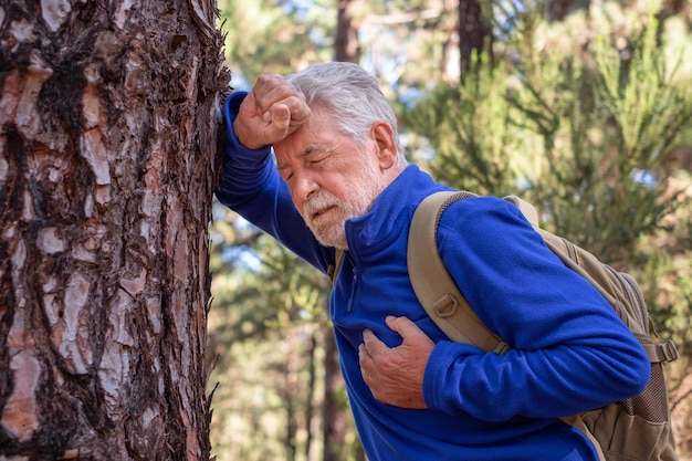 Un homme âgé aux cheveux blancs en randonnée dans les montagnes s'appuie contre un tronc d'arbre pour reprendre son souffle en plaçant une main sur son cœur Le grand-père aîné ne se sent pas bien