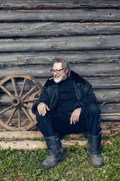 Un homme âgé avec une barbe se tient dans le village près d'un hangar en bois dans un manteau en peau de mouton et des lunettes