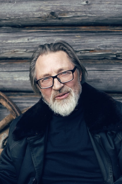 Un homme âgé avec une barbe se tient dans le village près d'un hangar en bois dans un manteau en peau de mouton et des lunettes