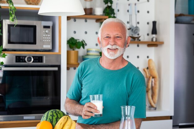 Homme âgé buvant un verre de lait avec un visage heureux debout et souriant Bel homme âgé buvant un verre de lait frais dans la cuisine