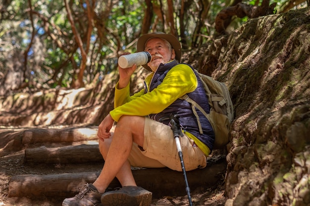 Homme âgé caucasien souriant assis sur le chemin forestier pour se reposer de l'eau potable de la fiole