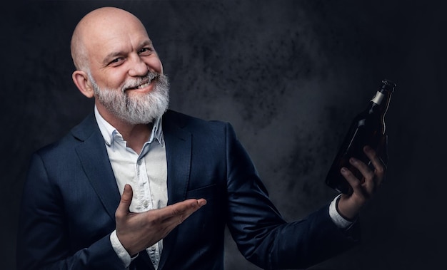 Homme âgé chauve avec barbe grise et bouteille posant sur fond sombre.