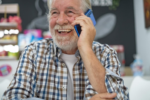 Homme âgé en chemise à carreaux assis à la table d'un café intérieur utilisant un téléphone portable parlant et riant