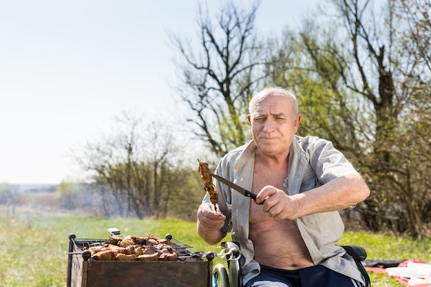 Un homme âgé avec une chemise déboutonnée et assis sur son fauteuil roulant tenant de la viande grillée sur un bâton et un couteau et regardant la caméra tout en faisant des grillades dans le parc.