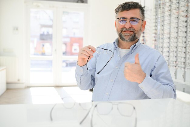 Un homme âgé choisissant des lunettes dans un magasin d'optique essaie des montures de lunettes pour décider de la parfaite