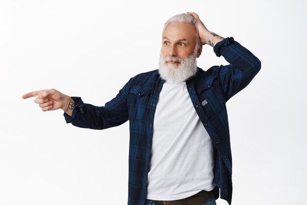 Un Homme âgé Confus Pointe Le Doigt Vers Quelque Chose De Surprenant, Se Gratte La Tête Et Regarde Avec Admiration, Regarde Quelque Chose De Cool, Regarde Là Un Geste, Debout Sur Un Mur Blanc