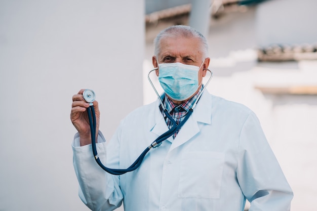 Un homme âgé dans une blouse blanche et un masque médical sur son visage montre un stéthoscope pour examiner le travail des poumons et du cœur des patients