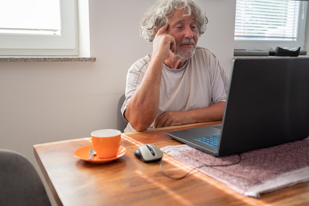 Homme âgé, grand-père, aux cheveux gris bouclés, assis à table à manger, parlant avec sa famille par appel vidéo sur ordinateur portable.