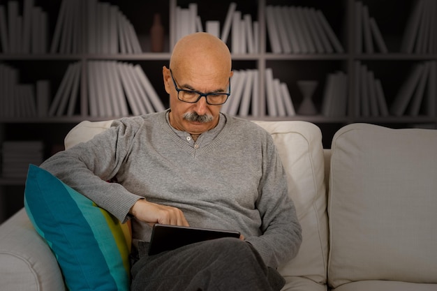 Homme âgé lisant dans une tablette numérique assis sur le canapé dans le salon de la maison