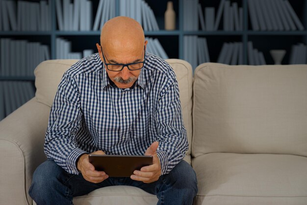 Homme âgé lisant dans une tablette numérique assis sur le canapé dans le salon de la maison