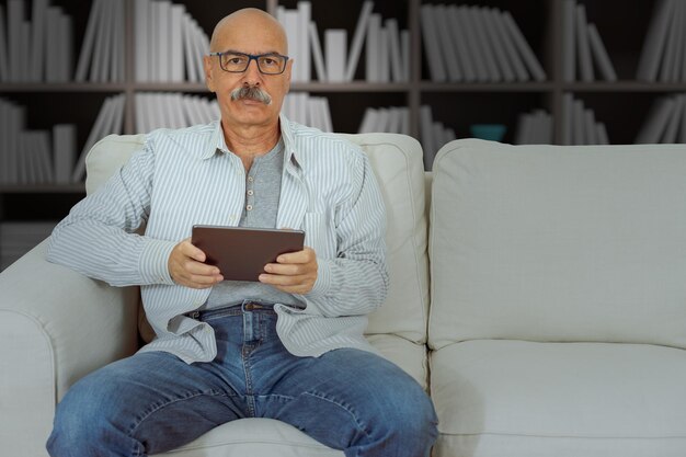 Homme âgé lisant dans une tablette numérique assis sur le canapé dans le salon de la maison