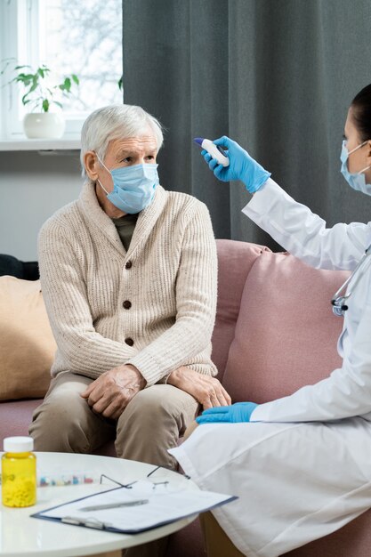 Homme âgé malade regardant une jeune femme médecin en blouse blanche à l'aide d'un thermomètre sans contact tout en mesurant la température de son corps