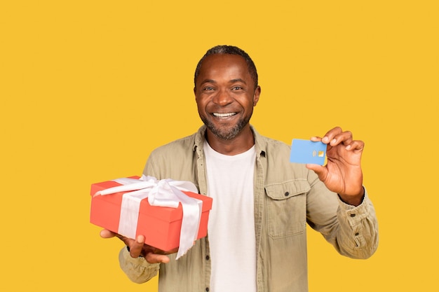 Un homme d'âge moyen afro-américain joyeux montre une boîte-cadeau et une carte de crédit recommande les services bancaires