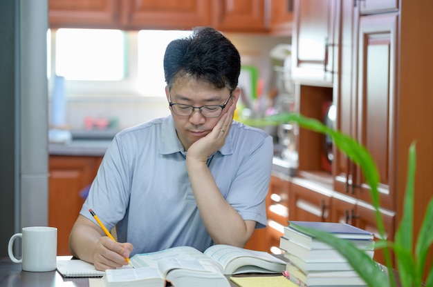 Homme d'âge moyen asiatique assis au bureau à la maison, lisant un livre et étudiant.