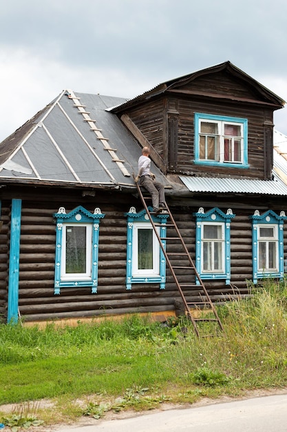 Homme d'âge moyen assis sur une échelle en métal réparant le toit de la vieille maison brune en bois sculptée de fenêtres