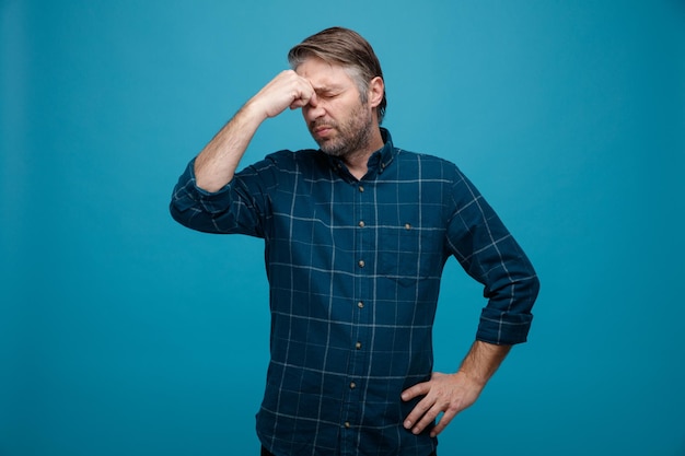 Homme D'âge Moyen Aux Cheveux Gris En Chemise De Couleur Foncée à La Confusion Et Déçu Tenant Les Doigts Entre Les Yeux Debout Sur Fond Bleu