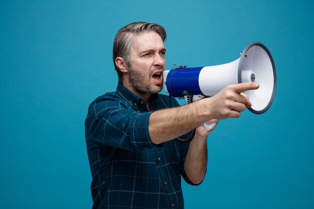 Homme d'âge moyen aux cheveux gris en chemise de couleur foncée criant dans un mégaphone pointant avec l'index vers quelque chose debout sur fond bleu