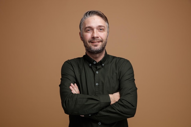 Homme d'âge moyen aux cheveux gris en chemise de couleur foncée regardant la caméra souriant confiant croisant les mains sur sa poitrine debout sur fond marron