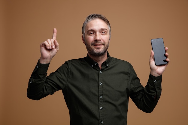 Homme d'âge moyen aux cheveux gris en chemise de couleur foncée tenant un smartphone pointant avec l'index vers le haut heureux et positif regardant la caméra souriant debout sur fond marron