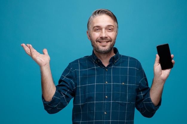 homme d'âge moyen aux cheveux gris en chemise de couleur foncée tenant un smartphone regardant la caméra heureux et souriant levant le bras debout sur fond bleu