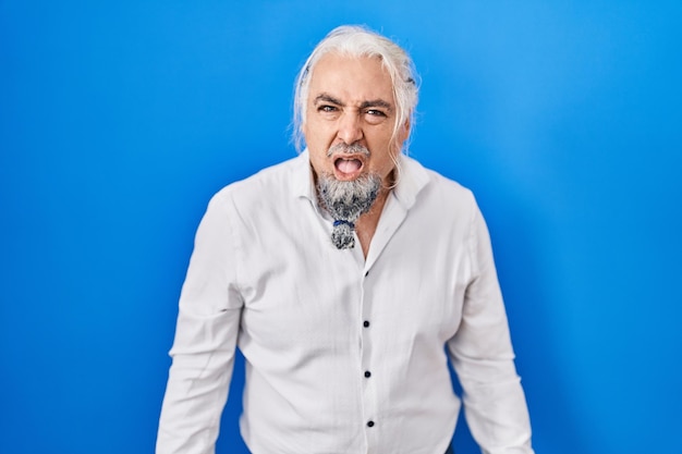 Homme d'âge moyen aux cheveux gris debout sur fond bleu en face de choc, à l'air sceptique et sarcastique, surpris avec la bouche ouverte