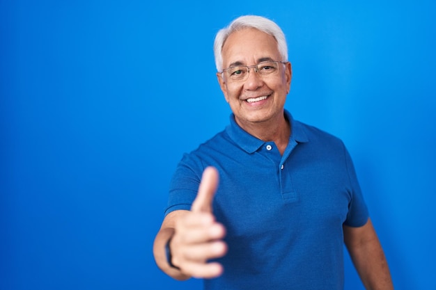 Homme d'âge moyen aux cheveux gris debout sur fond bleu souriant amical offrant une poignée de main comme salutation et accueillant une entreprise prospère