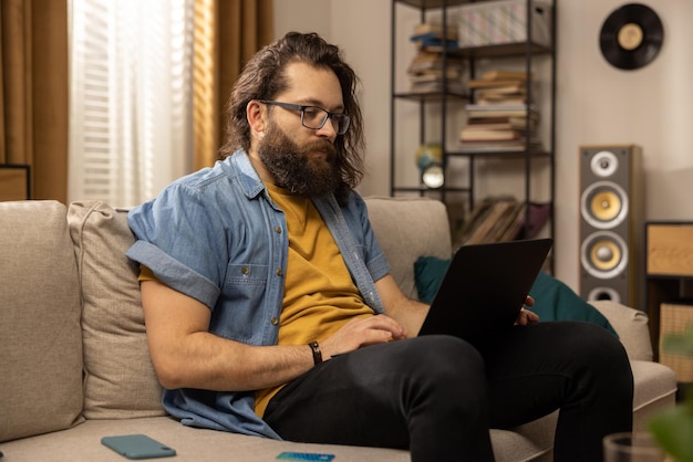 Un homme d'âge moyen avec une barbe utilise un ordinateur portable assis sur le canapé dans le salon un étudiant