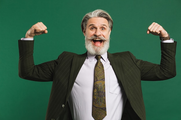 Homme d'âge moyen barbu en costume montre les biceps et sourit émotionnellement sur le mur vert