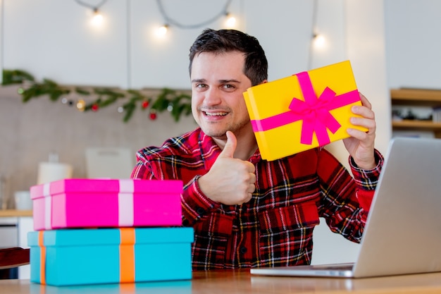 Homme d'âge moyen blanc en t-shirt rouge tenant une boîte-cadeau et à la recherche de ventes sur Internet via un ordinateur portable au moment de Noël.