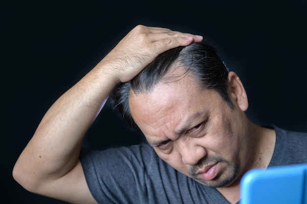 Un homme d'âge moyen brille dans le miroir et s'inquiète de la perte de cheveux ou des cheveux gris isolés sur fond noir Concept de soins de santé