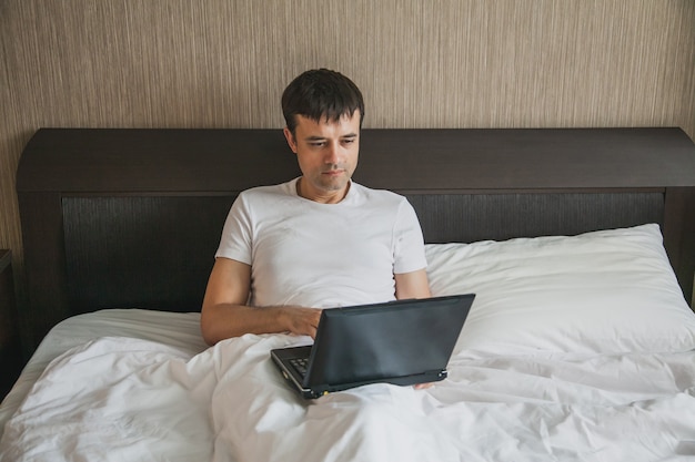 Un homme d'âge moyen est assis dans son lit dans sa chambre et travaille sur un ordinateur portable. Le concept de télétravail