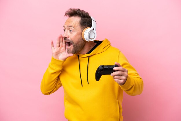 Homme d'âge moyen jouant avec un contrôleur de jeu vidéo isolé sur fond rose criant avec la bouche grande ouverte sur le côté