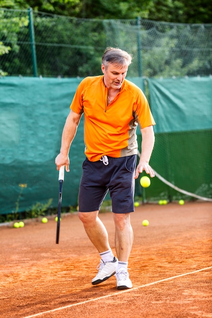 Un homme d'âge moyen joue au tennis sur un court avec une surface en terre naturelle sur une journée d'été ensoleillée.