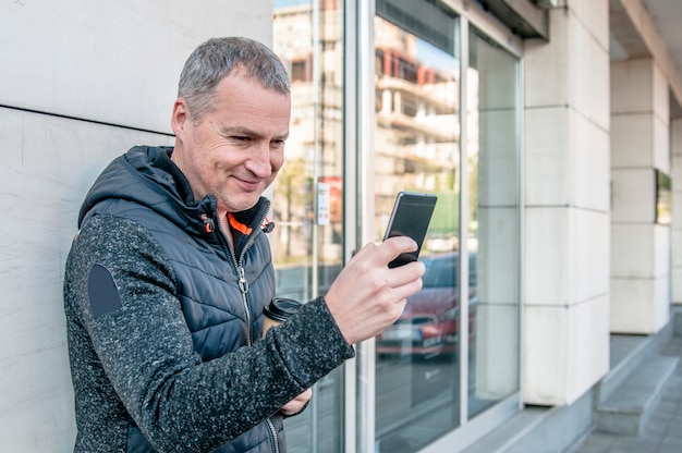 Un homme d&#39;âge moyen marchant à côté du bâtiment de bureau tout en utilisant son smartphone