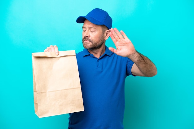 Homme d'âge moyen prenant un sac de plats à emporter isolé sur fond bleu faisant un geste d'arrêt et déçu