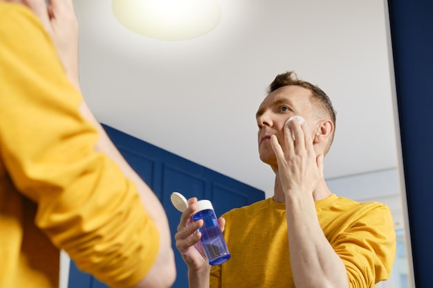 Homme d'âge moyen regardant dans le miroir et nettoyant le visage avec un coton