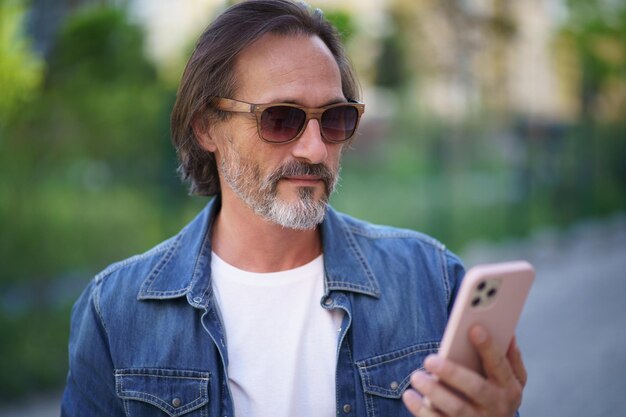 Homme d'âge moyen regardant un smartphone debout dans la rue tout en profitant de son temps libre hors travail