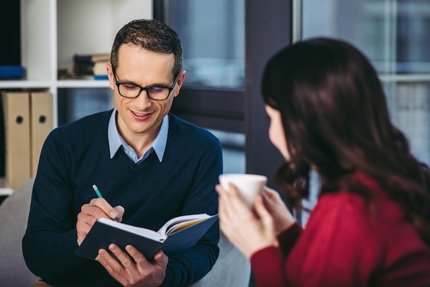 Homme d'âge moyen souriant prenant des notes dans un cahier tandis qu'une femme buvant du café et le regardant