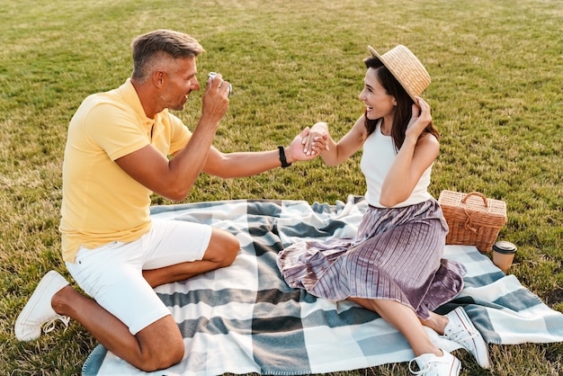 Photo homme d'âge moyen souriant prenant une photo d'une belle femme sur un appareil photo rétro alors qu'il était assis sur l'herbe dans le parc d'été