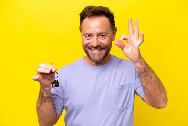 Homme d'âge moyen tenant les clés de la maison isolé sur fond jaune montrant le signe ok avec les doigts