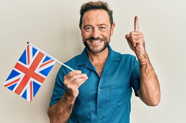 Homme d'âge moyen tenant le drapeau du royaume-uni souriant avec une idée ou une question pointant du doigt avec un visage heureux, numéro un