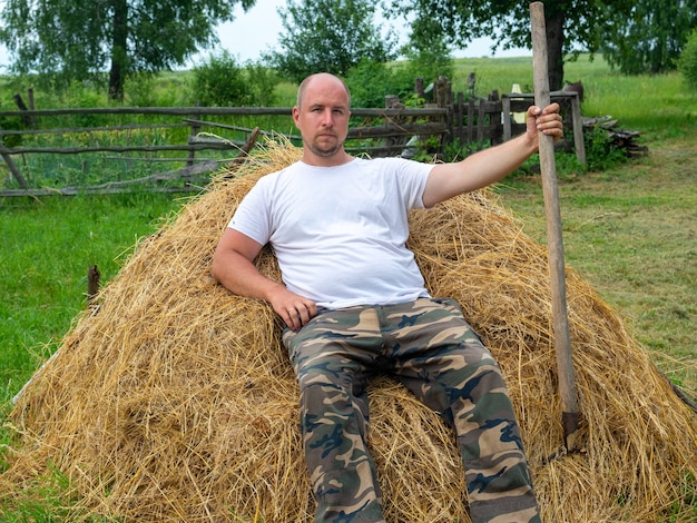 Un homme d'âge moyen vêtu d'un pantalon et d'un T-shirt est assis sur une pile de foin jaune et tient une fourche à la main. Le concept des zones rurales et des travailleurs agricoles