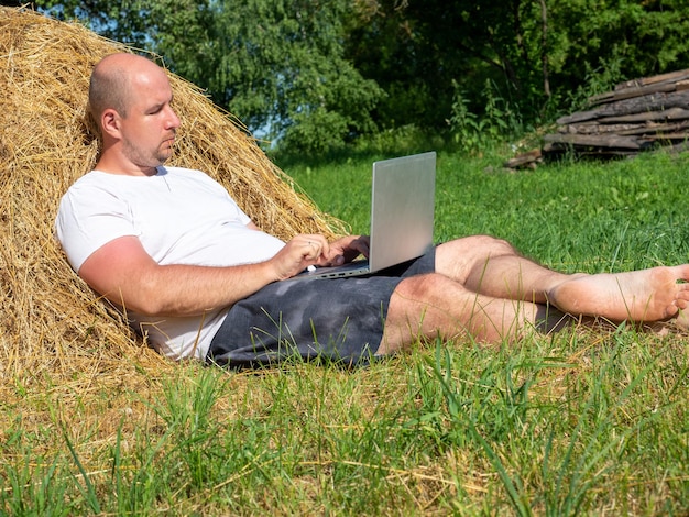 Un homme d'âge moyen vêtu d'un t-shirt et d'un short est allongé sur une botte de foin jaune avec un ordinateur portable gris dans ses mains Formation au travail à distance Zone rurale