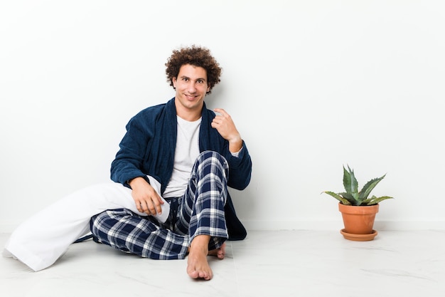 Homme d'âge mûr portant un pyjama assis sur le sol de la maison pointant du doigt vers vous comme si vous vous invitiez à vous rapprocher.