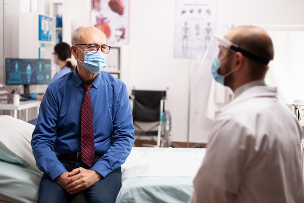 Homme âgé parlant avec un médecin du traitement assis sur un masque de protection du lit comme précaution de sécurité Crise sanitaire mondiale, système médical pendant la pandémie de coronavirus, patient âgé malade en privé cl