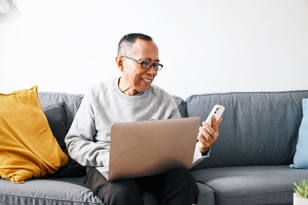 Homme âgé portant des lunettes utilisant un téléphone portable alors qu'il est assis sur son canapé confortable avec un ordinateur portable sur les genoux