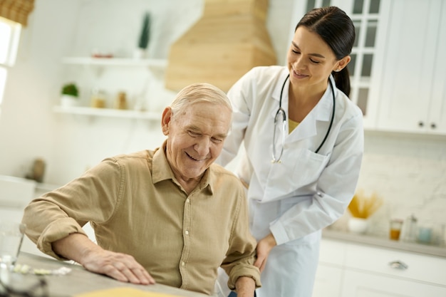 Homme âgé positif souriant en se levant de la chaise avec l'aide d'un jeune médecin attentif