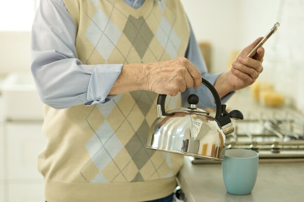 Homme âgé regardant et smartphone et versant de l'eau de la bouilloire