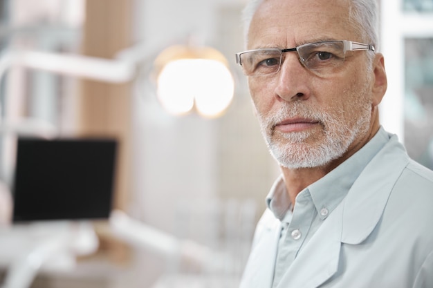 Homme âgé réservé portant des lunettes et une blouse de laboratoire debout dans le bureau du dentiste et regardant la caméra